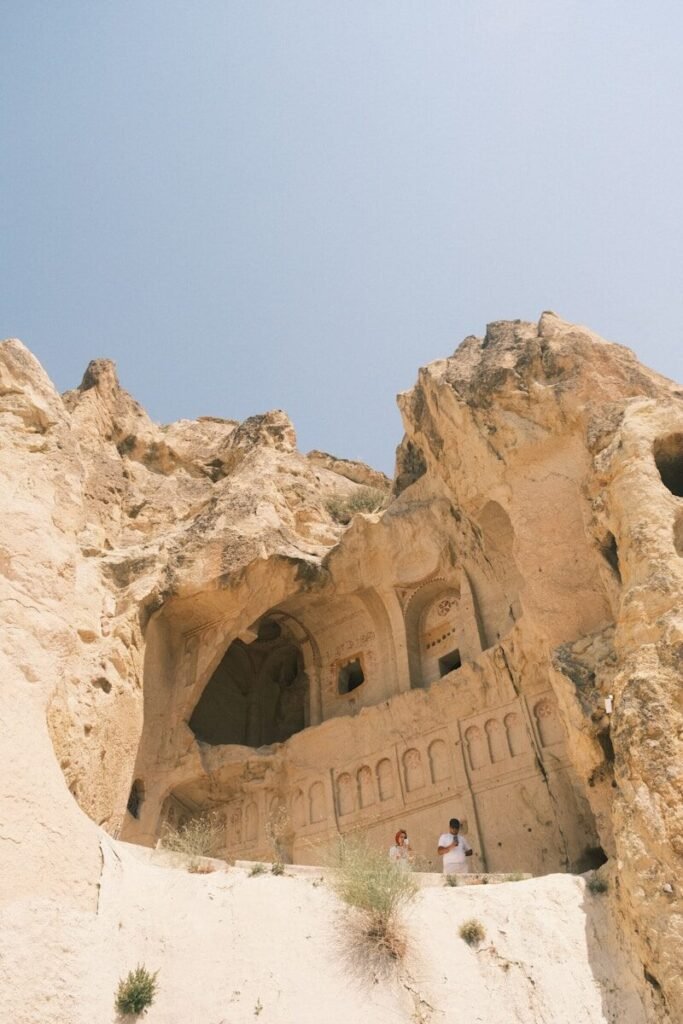 Two people stand near ancient cave dwellings in rock formations.